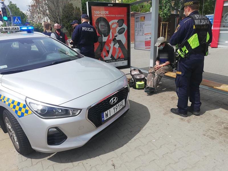 Ujęty mężczyzna siedzi na przystanku. Obok, przy zaparkowanym radiowozie, stoi strażnik miejski. W&nbsp;tle policjanci rozmawiają ze świadkami