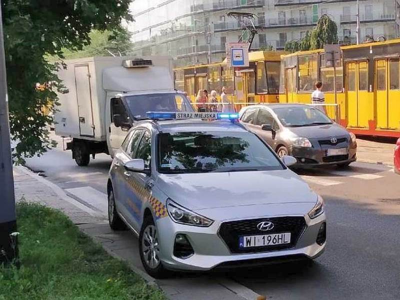 Radiowóz straży miejskiej stojący na poboczu jezdni. W&nbsp;tle przystanek ze stojącym na nim tramwajem.