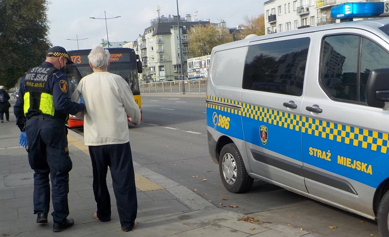 Zdjęcie z interwencji: 64-latek ubrany w piżamę prowadzony pod rękę przez strażnika miejskiego w kierunku radiowozu straży miejskiej.