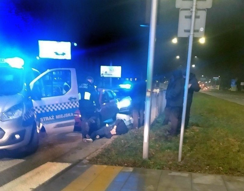 Zdjęcie z&nbsp;interwencji: po lewej stronie obrazu stojący radiowóz straży miejskiej z&nbsp;włączonymi sygnałami świetlnymi, za nim zatrzymany samochód. Obok leżący na poboczu mężczyzna zakuty w&nbsp;kajdanki. Nad nim stoją strażnicy miejscy.