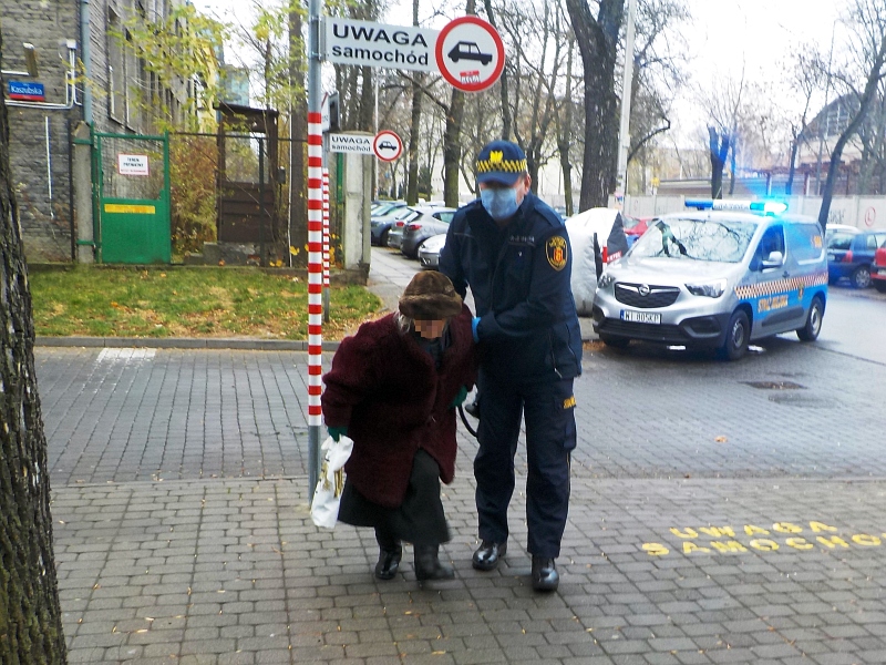 Zdjęcie z interwencji: strażnik miejski prowadzący ubraną w ciemnoczerwony płaszcz starszą kobietę.