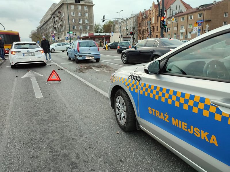 Zdjęcie z&nbsp;interwencji: na pierwszym planie radiowóz Straży Miejskiej, za nim, na jezdni, stoi trójkąt ostrzegawczy. Dalej widać dwa samochody, które wzięły udział w&nbsp;kolizji- błękitny po prawej, biały po lewej. Przy nim stoją dwie osoby.