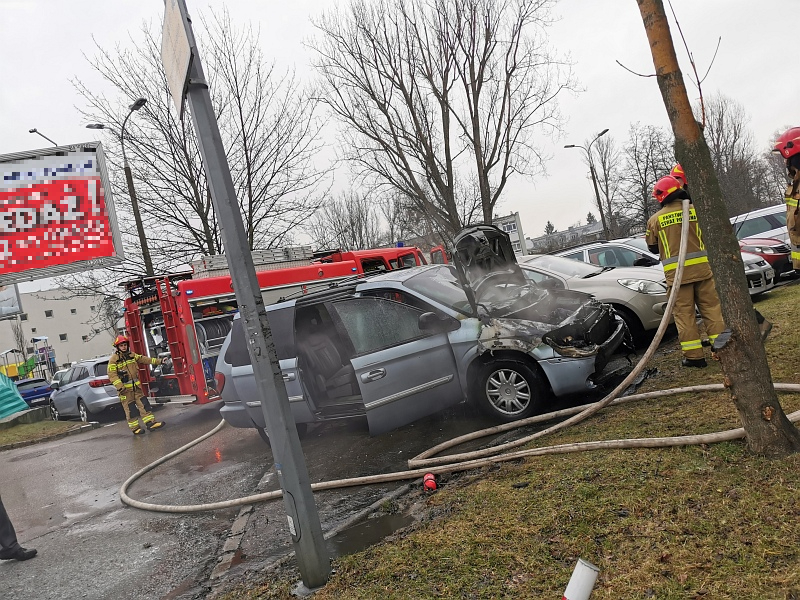 Zdjęcie z interwencji: w środku kadru srebrny chrysler z widocznymi śladami spalenia, obok niego dogaszający pożar strażacy. W tle samochód strażacki oraz zaparkowane samochody.