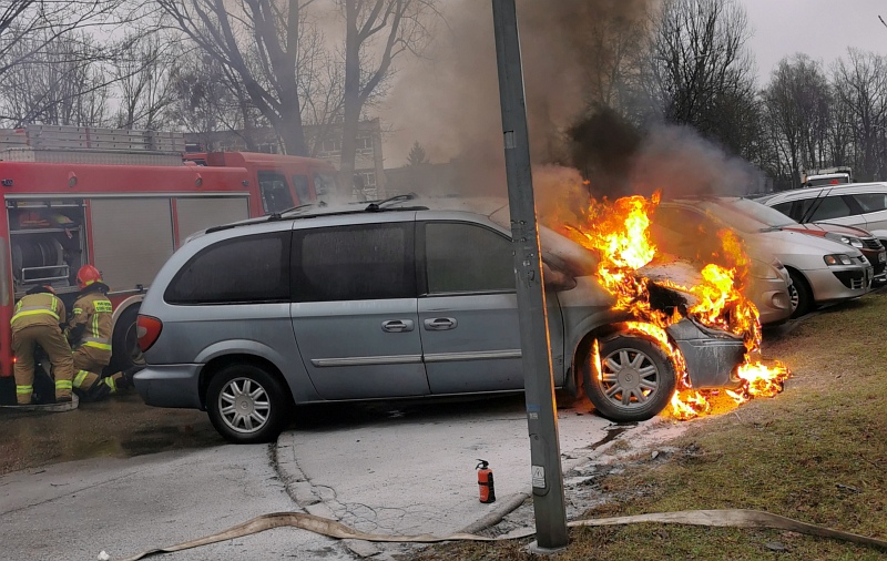 Zdjęcie z interwencji: srebrny chrysler z płomieniami wydobywającymi się spod maski. Za nim strażacy przygotowujący się do akcji gaśniczej. Obok, na asfalcie, stoi gaśnica, którą wcześniej strażnicy miejscy usiłowali stłumić pożar.