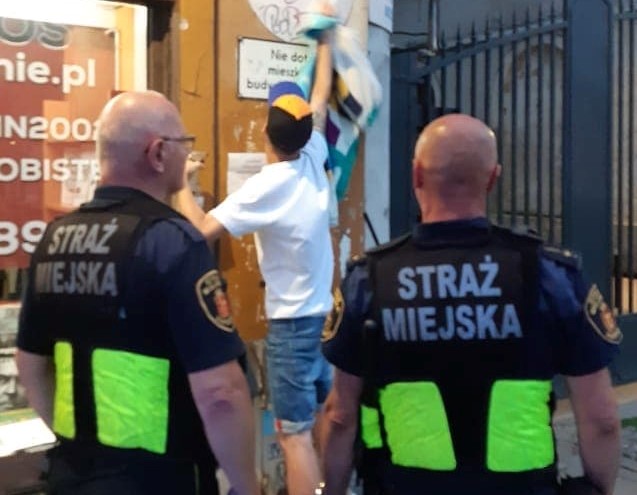 Młody mężczyzna ściera ze ściany namazane wcześniej graffiti. Przywracanie poprzedniego stanu elewacji nadzorują dwaj strażnicy miejscy.