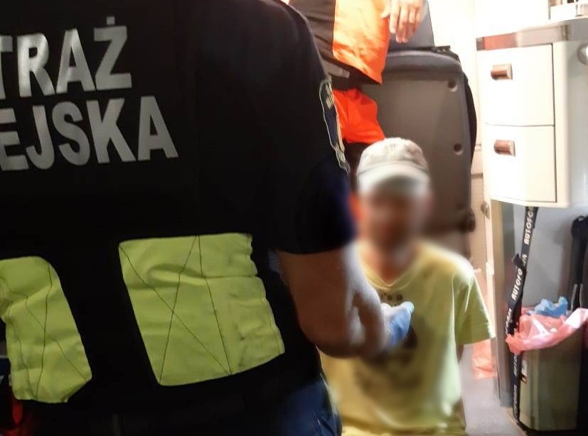 Mężczyzna w żółtym T-shircie siedzi na wejściu do ambulansu, przed którym stoi strażnik miejski (widać plecy).