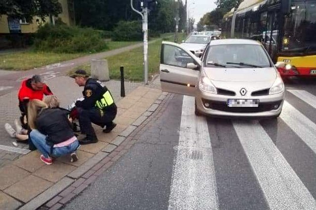 Na jezdni stoi beżowe renault. Obok, na chodniku, przy przejściu dla pieszych, leży poszkodowana kobieta. Nad nią dwoje ludzi oraz strażnik miejski udzielający pierwszej pomocy.