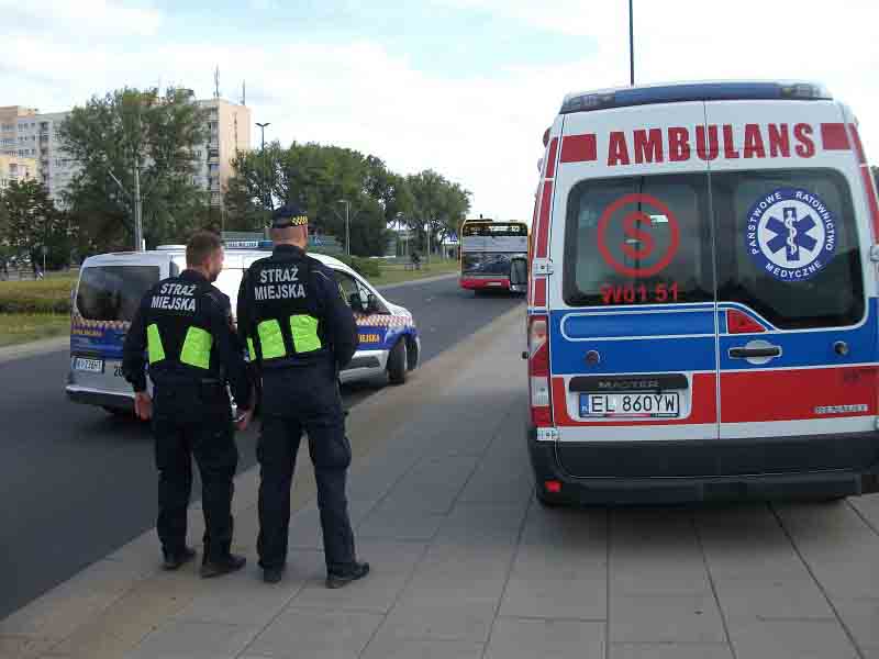 Strażnicy miejscy stojący obok karetki pogotowia ratunkowego.