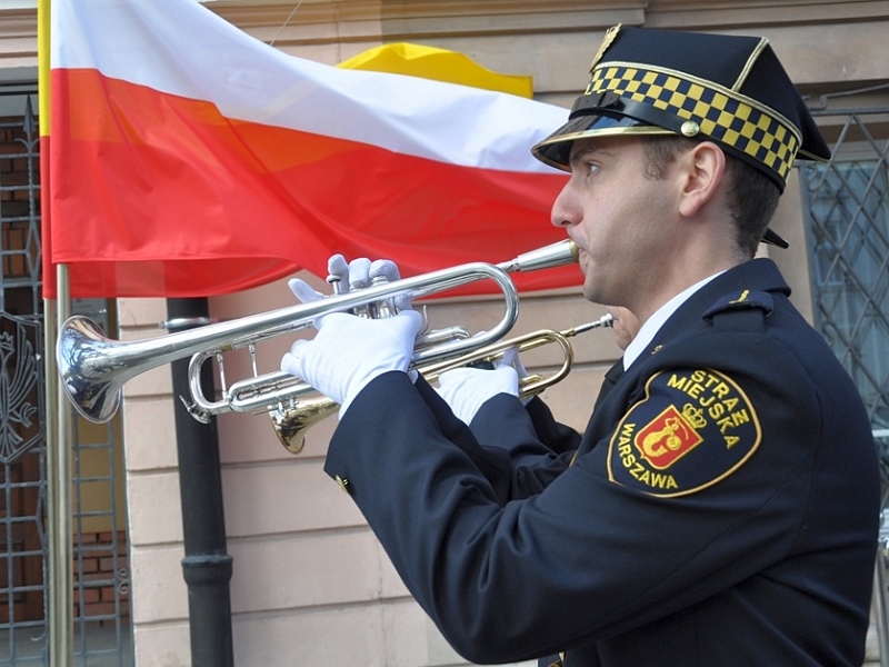 Hejnalista straży miejskiej grający na trąbce. W tle flagi Polski i Warszawy.