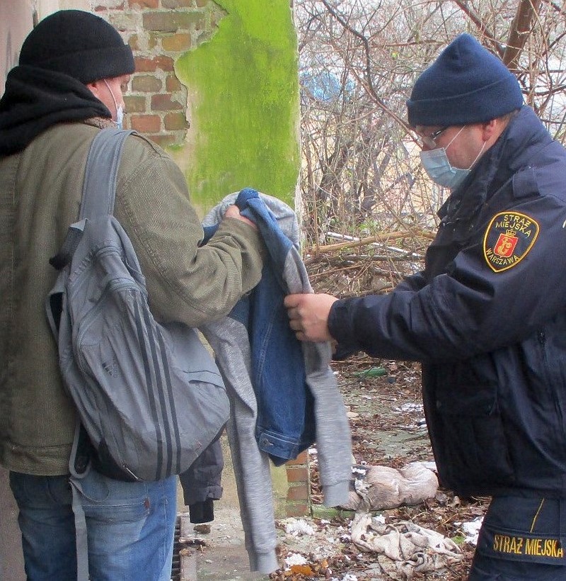 Zdjęcie ilustracyjne: stojący po prawej stronie kadru strażnik miejski podaje bezdomnemu, stojącemu z lewej, bluzę. W tle opuszczony budynek.