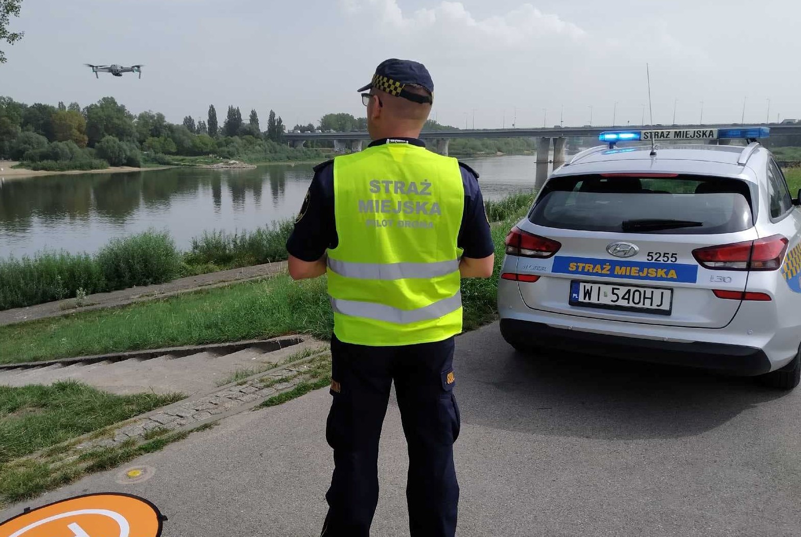 Strażnik miejski w trakcie oblotu dronem okolic jeziorka Gocławskiego. Dron w locie, strażnik z urządzeniem sterującym i radiowóz straży miejskiej nad wodą.