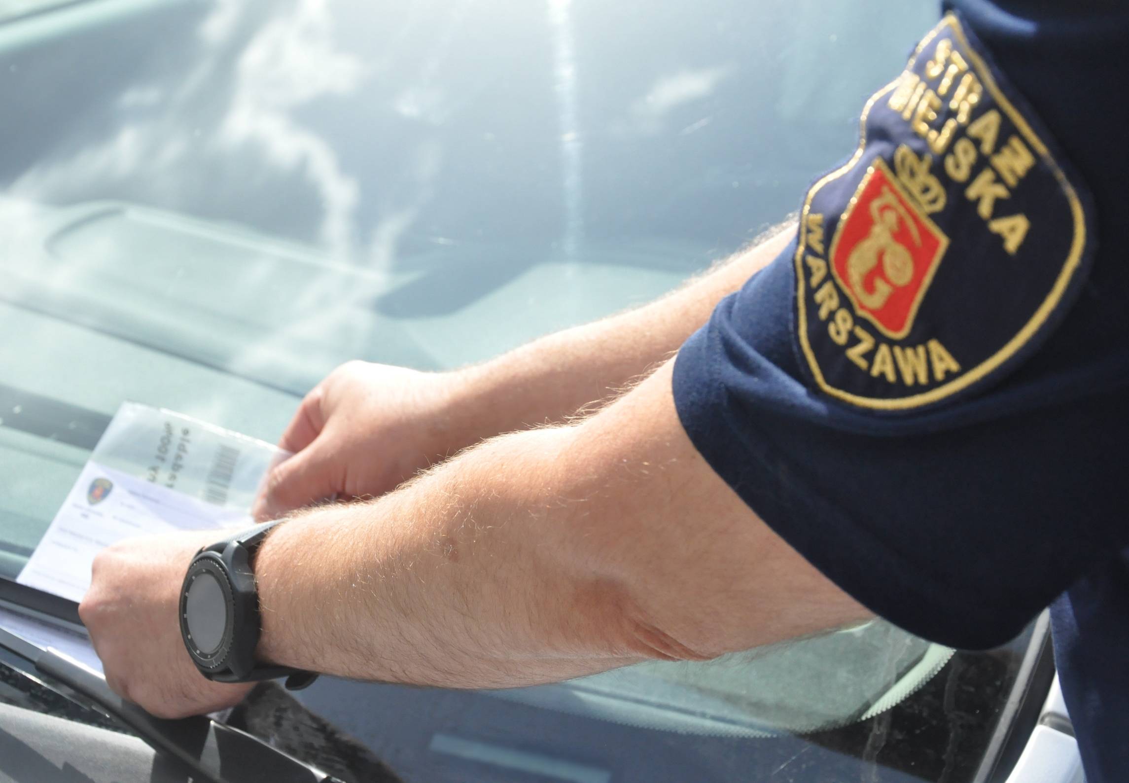 Strażnik miejski umieszcza wezwanie za wycieraczką samochodową.