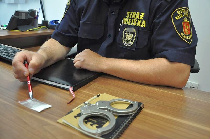 Siedzący za biurkiem strażnik wskazuje długopisem na foliową torebkę z białym proszkiem. Obok niej, na biurku, leżą kajdanki.