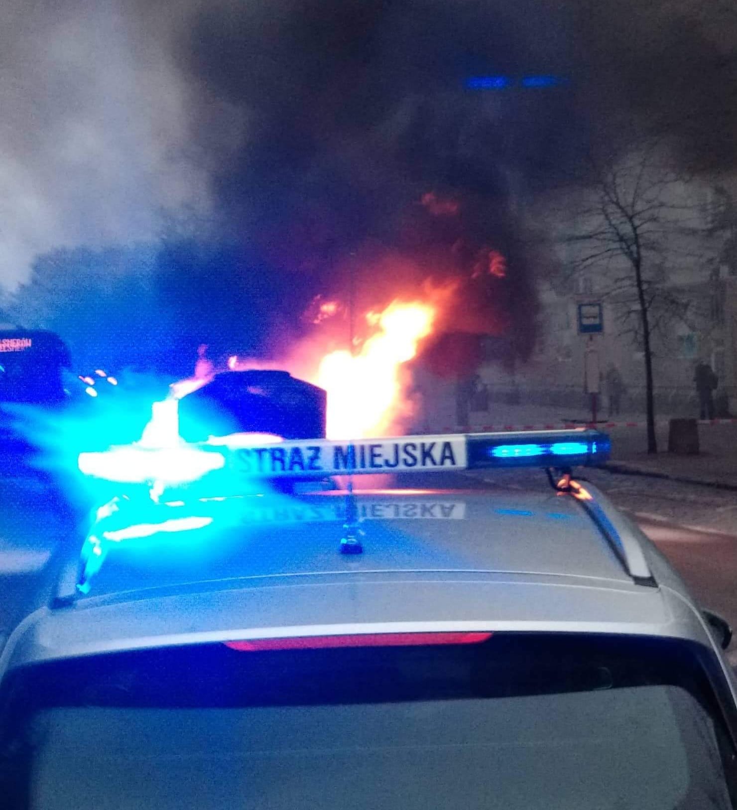 Radiowóz straży miejskiej z włączonymi sygnałami świetlnymi. W tle płonący samochód. Zdjęcie ilustracyjne.