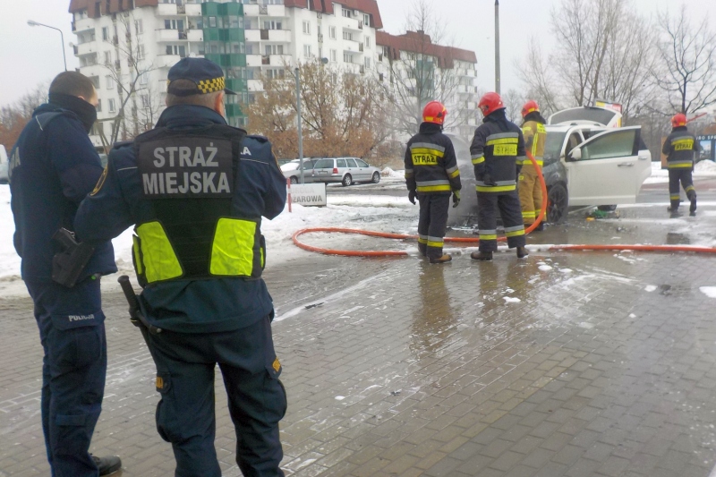 Strażnicy zabezpieczają miejsce pożaru. W&nbsp;tle strażacy dogaszają samochód.