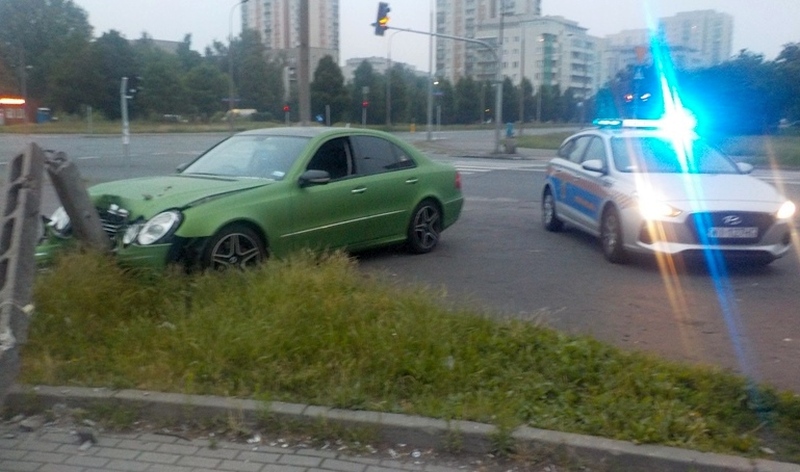 Zielony mercedes wbity w słup latarni. Na prawo od niego radiowóz straży miejskiej z włączonymi światłami sygnalizacyjnymi.