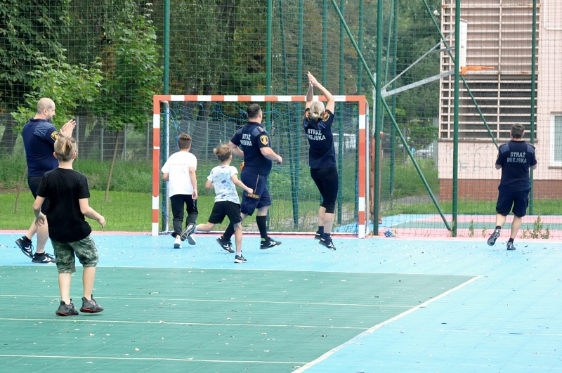 Boisko piłkarskie, przed bramką trzech chłopców i&nbsp;czworo strażników. Jedna z&nbsp;nich w&nbsp;triumfalnym geście unosi ręce do góry ciesząc się po zdobytej bramce.