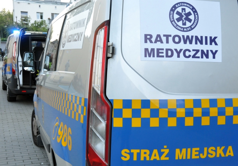 Na pierwszym planie widoczny tył radiowozu straży miejskiej z napisem "Ratownik medyczny". w tle taki sam radiowóz z otwartymi tylnymi drzwiami i włączonymi światłami sygnalizacyjnymi.