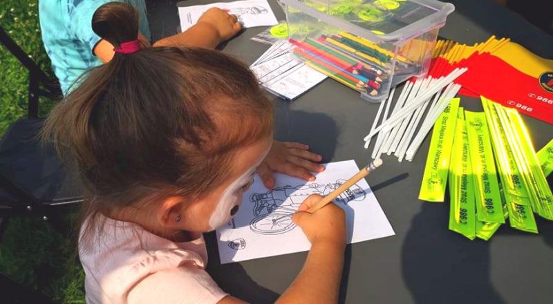 Dziewczynka z&nbsp;pomalowaną buzią koloruje obrazek przy stanowisku Straży Miejskiej. Na stoliku leżą flagi Warszawy, opaski odblaskowe i&nbsp;kredki.