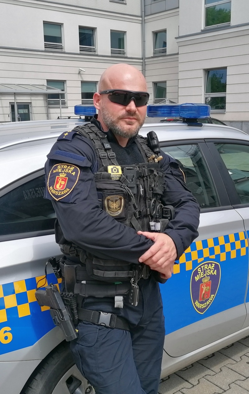 Uczestnik zawodów, inspektor Łukasz Capiga, stoi oparty o&nbsp;radiowóz straży miejskiej.