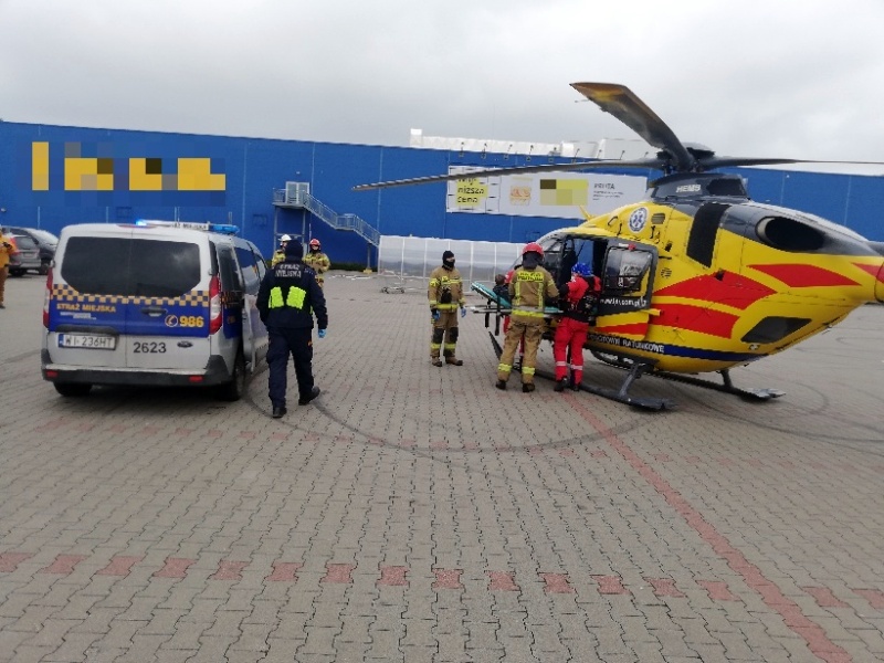 Zdjęcie z interwencji: parking centrum handlowego. Z lewej strony kadru stoi radiowóz straży miejskiej, obok- strażnik miejski. Po prawej stronie żółto-czerwony śmigłowiec Lotniczego Pogotowia Ratunkowego, a przy nim ratownicy i strażacy.