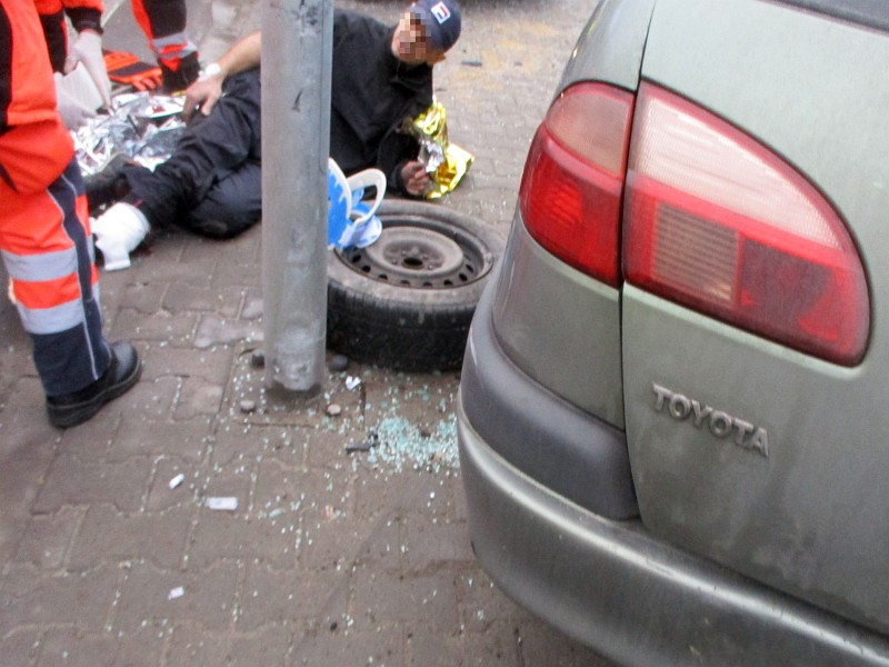 Zdjęcie z interwencji: na pierwszym planie tył uszkodzonej toyoty, w tle, obok koła zapasowego, leży mężczyzna z ranną nogą opatrywany przez ratowników medycznych.