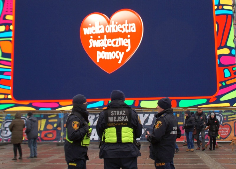 Zdjęcie z&nbsp;wydarzenia: pod telebimem z&nbsp;logo Wielkiej Orkiestry Świątecznej Pomocy stoi trójka strażników miejskich.
