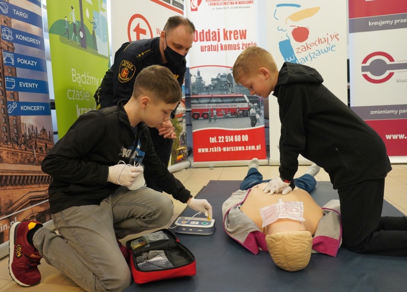 Zdjęcie z&nbsp;trzeciego etapu konkursu: dwóch chłopców wykonuje masaż serca na fantomie . Przygląda im się strażnik miejski.