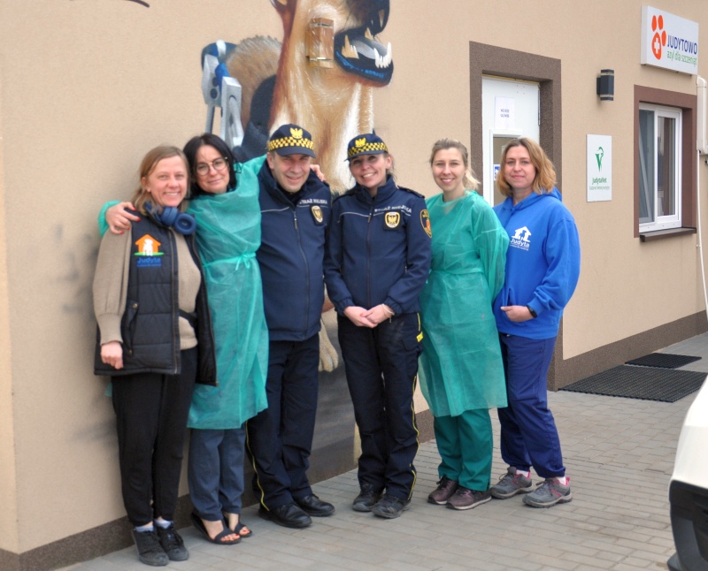 Zdjęcie grupowe: dwoje strażników miejskich stojących w&nbsp;otoczeniu pracownik schroniska na tle muralu z&nbsp;psem.