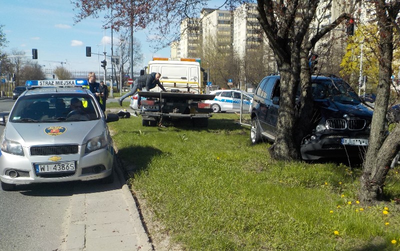 Zdjęcie z interwencji: z prawej strony czarne BMW zatrzymane na dwóch drzewkach, z lewej- stojący na jezdni radiowóz straży miejskiej. Między nimi laweta pomocy drogowej.