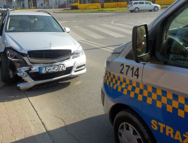 USzkodzony w kolizji mercedes stojący na poboczu jezdni. Przed nim, na pierwszym planie, radiowóz straży miejskiej.