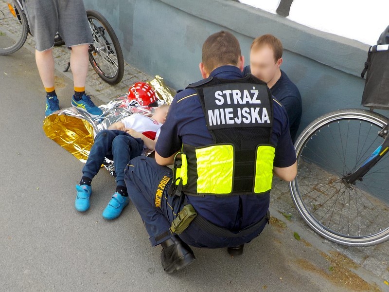 Strażnik miejski klęczący nad leżącym na kocu ratunkowym siedmiolatku. Obok chłopca siedzi jego ojciec, oparty o&nbsp;ścianę.