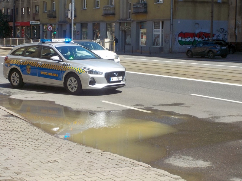 RAdiowóz straży miejskiej zabezpieczający miejsce awarii. Na jezdni i chodniku widoczne rozlewiska wody.