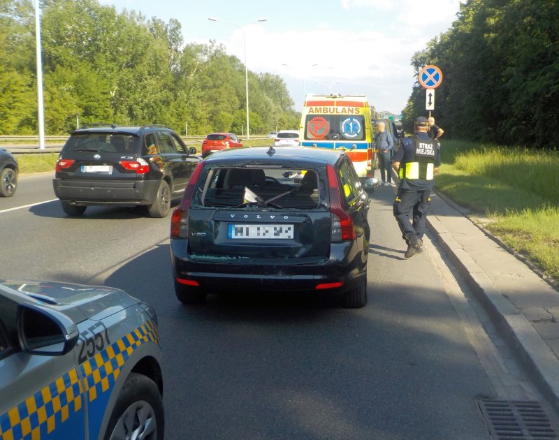 Czarne volvo uszkodzone w kolizji. Obok niego strażnik miejski, na dalszym planie karetka pogotowia.