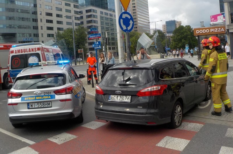 Samochód uszkodzony w wypadku stoi na ścieżce rowerowej, na rondzie ONZ. Obok zabezpieczający zdarzenie radiowóz Straży Miejskiej miasta stołecznego Warszawy . Dalej karetka pogotowia, strażacy i przechodnie.