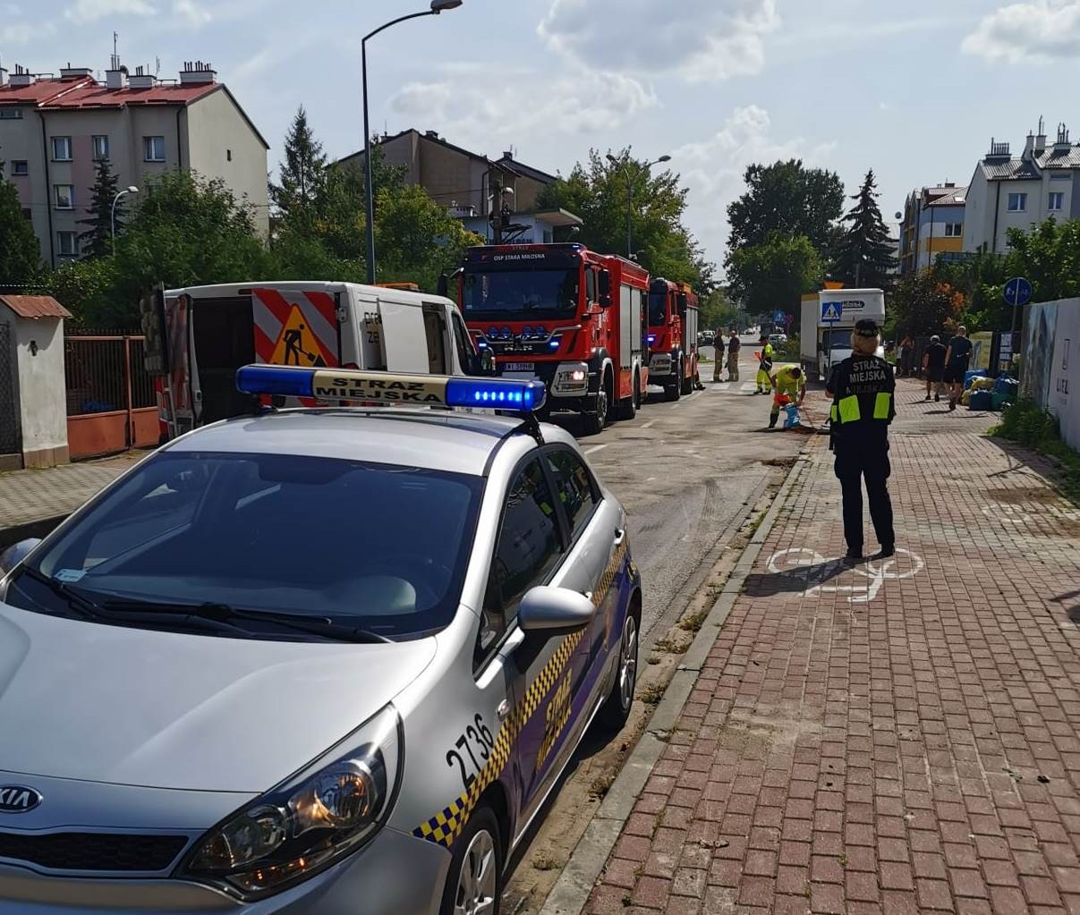 Strażnicy miejscy zabezpieczają pracę służby porządkowej i strażaków w miejscu zanieczyszczenia drogi olejem. Radiowóz straży miejskiej zastawiający przejazd niebezpiecznym pasem, strażniczka obserwująca użytkowników drogi, robotnicy i strażacy neutralizujący niebezpieczną plamę.