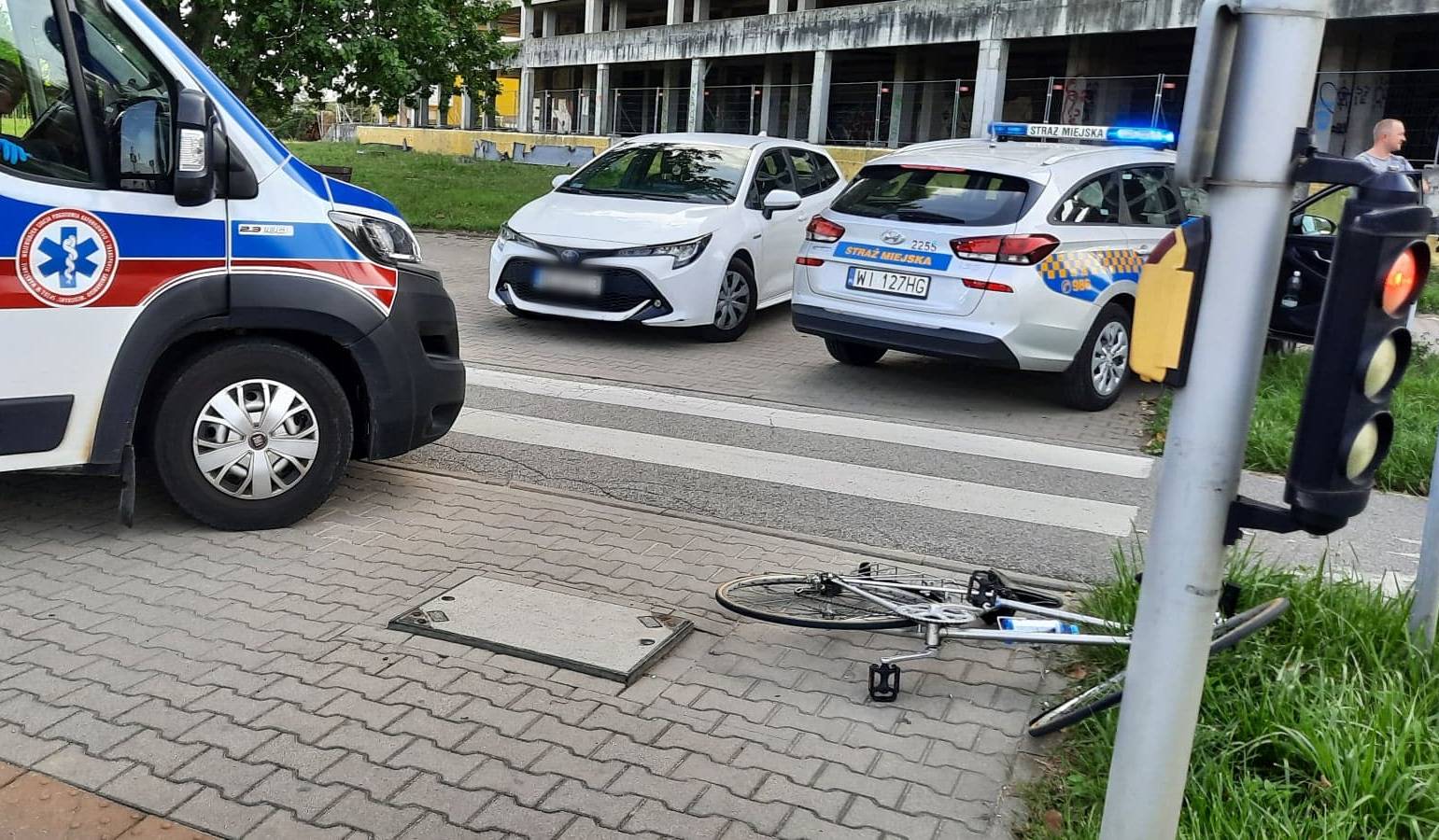 Miejsce wypadku. Rower leżący na chodniku. Obok karetka pogotowia. Z tyłu radiowóz straży miejskiej i samochód, który brał udział w zdarzeniu.