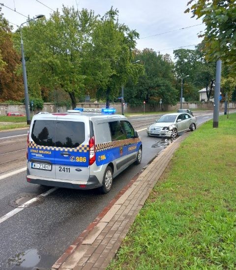 Radiowóz straży miejskiej zabezpieczający miejsce zdarzenia. W tle uszkodzona w kolizji toyota.