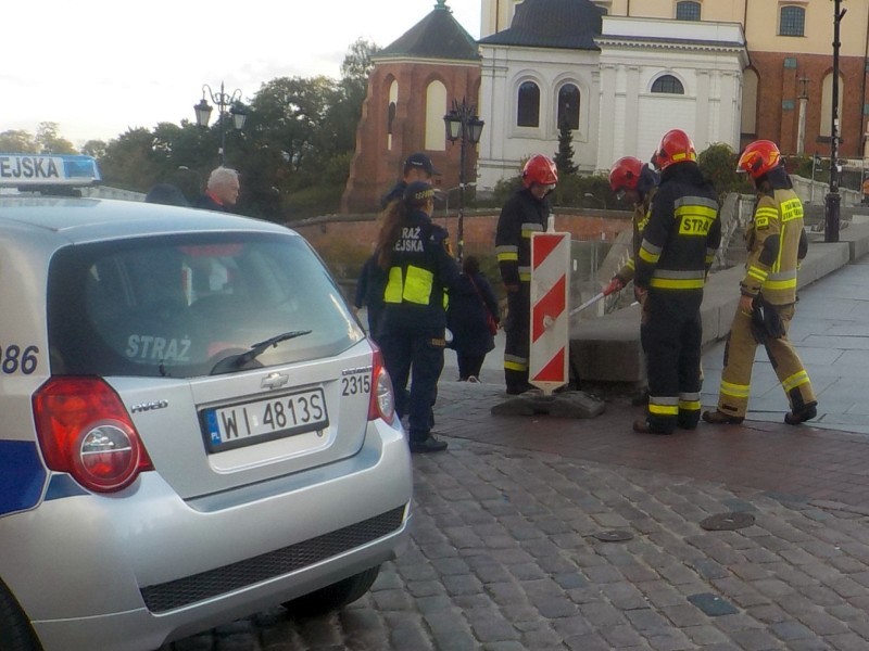 Na pierwszym planie radiowóz straży miejskiej. Za nim strażnicy zabezpieczający miejsce zdarzenia, obok strażacy gaszący płonące przyłącze po latarni ulicznej.