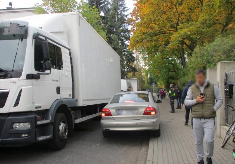 Zatarasowana ulica, w wolnym miejscu nie mieści się samochód osobowy. Na chodniku stoi strażnik miejski.